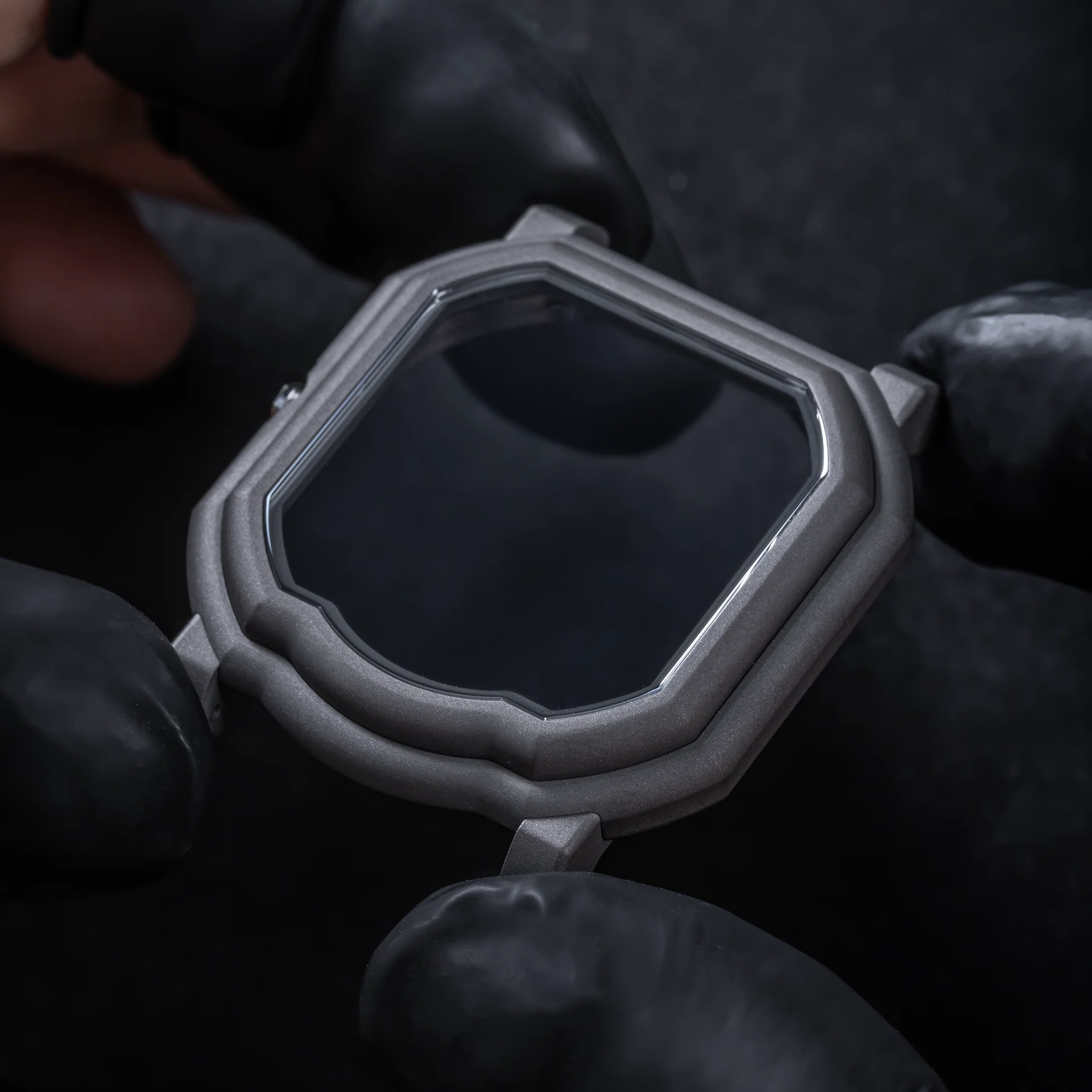 The bare Darkblast titanium Maestro case held in gloved hands, showing the stepped bezel and the curved indentation at six o'clock